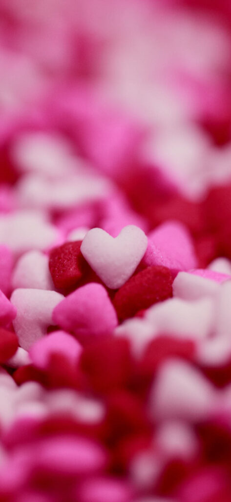 A close-up image of numerous small, heart-shaped candies in various shades of pink and red, with one white heart prominently in focus amidst them.