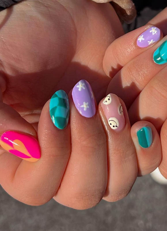 A close-up of a hand with nails painted in various colorful designs including geometric patterns, floral prints, and cute faces.

