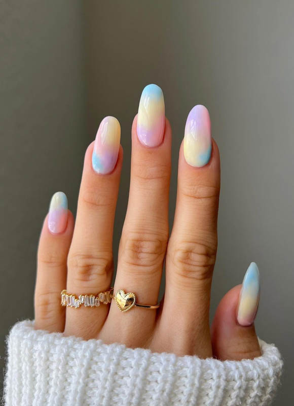 A hand with pastel gradient nails and rings spelling "Marry Me" on a grey background. Spring Nail Ideas For 2024.