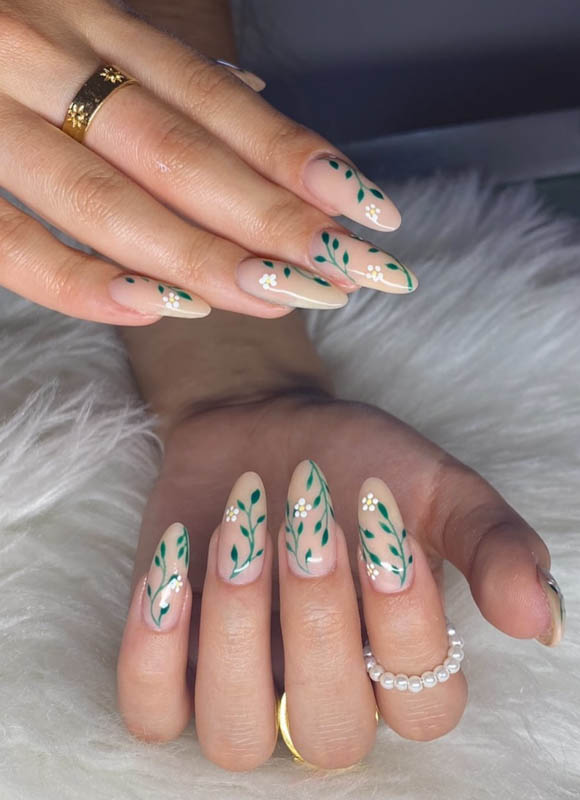 Spring Nail Designs For 2024. A close-up image of two hands with beautifully manicured nails, adorned with a delicate green leafy design, resting on a soft white surface. The person is wearing gold rings and a pearl bracelet.

