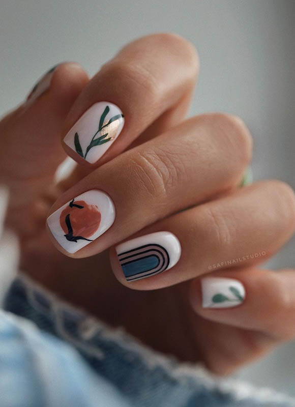 A close-up of a hand with nails artistically painted with various designs including a leafy branch, an abstract pattern, and a peach against a soft-focus background.

