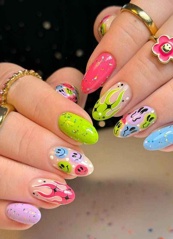 A close-up of a hand with nails painted in various bright colors and patterns, including faces, flames, and abstract designs, adorned with rings and a smiley face charm.

