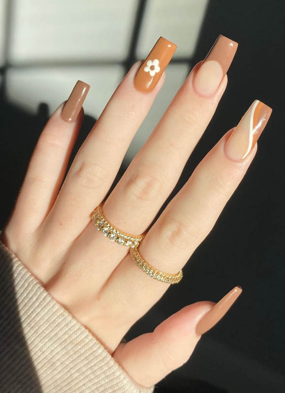 A hand with brown nails and two golden rings on a beige background. Spring Nail Ideas For 2024.