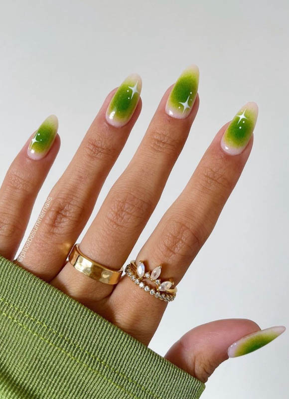 A hand with green and white ombre nails, wearing gold rings, against a white background.

