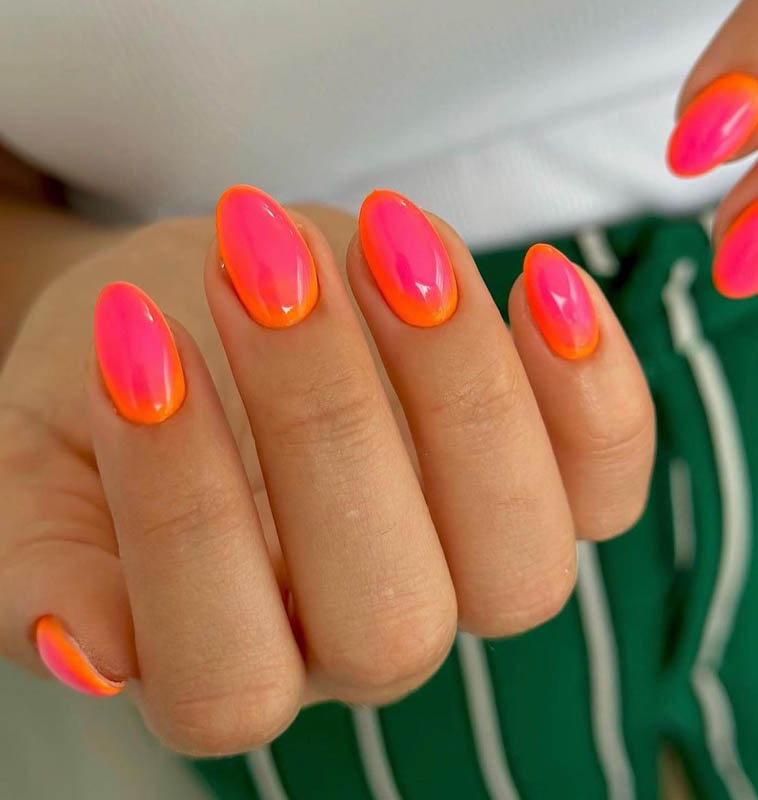 A hand with nails painted in a vibrant gradient of pink to orange, against a backdrop of white and green clothing. Spring Nail Ideas For 2024.