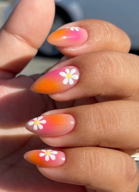 A close-up of a hand with nails painted in a gradient of pink to orange, adorned with small white flower decals. Spring Nail Ideas For 2024.