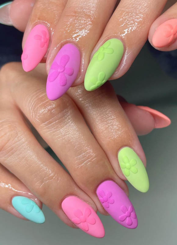 A close-up of hands with long, almond-shaped nails painted in pastel colors, each adorned with a raised floral design.

