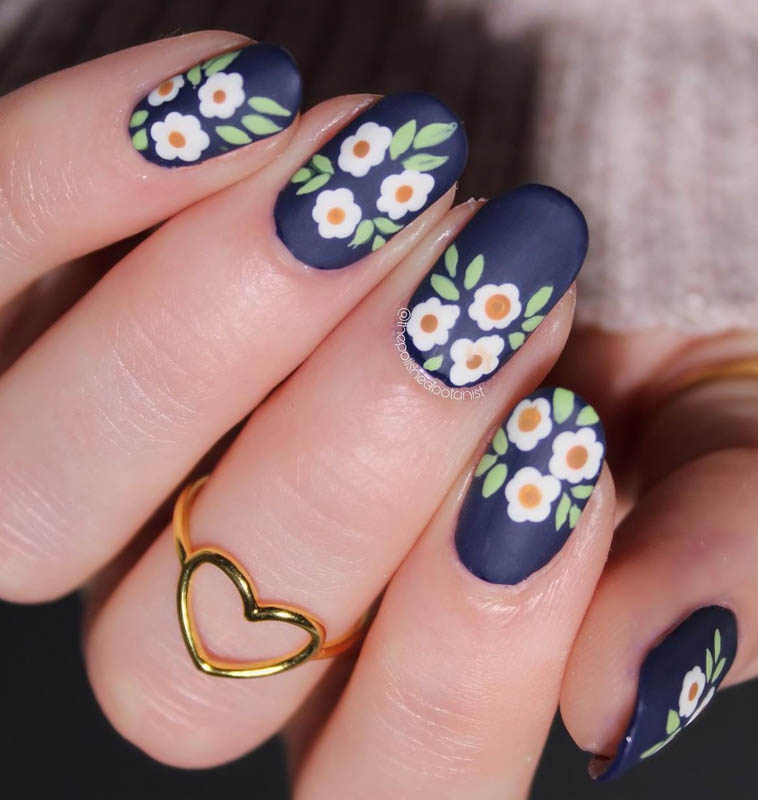 Spring Nail Designs For 2024. A hand with dark blue nails and white and orange floral designs, wearing a gold heart ring.

