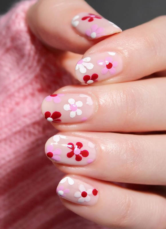 Spring Nail Designs For 2024. A close-up of a hand with nails artistically painted with white and red flowers against a soft pink background.

