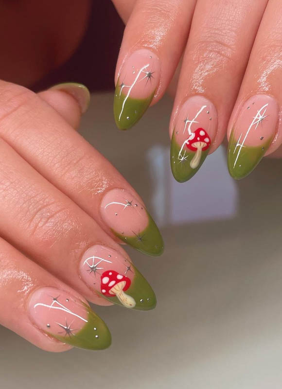 Spring Nail Designs For 2024. A close-up view of a hand with nails artistically painted in a gradient of green, adorned with delicate white starbursts and detailed red mushrooms with white spots.

