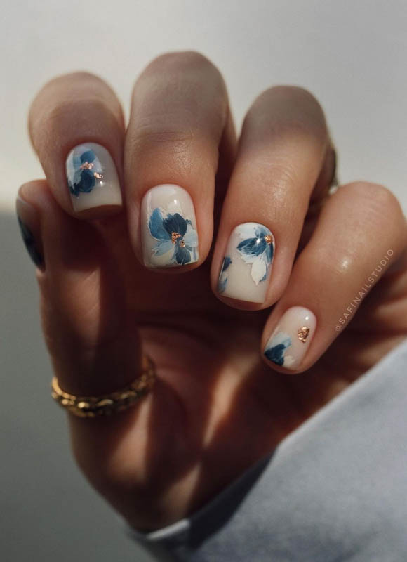 Spring Nail Designs For 2024. A close-up of a hand with nails artistically painted with delicate blue flowers against a soft white background, adorned with a gold ring on the pinky finger.

