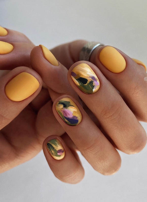 A hand with yellow nails and floral designs, wearing a ring, against a neutral background.

