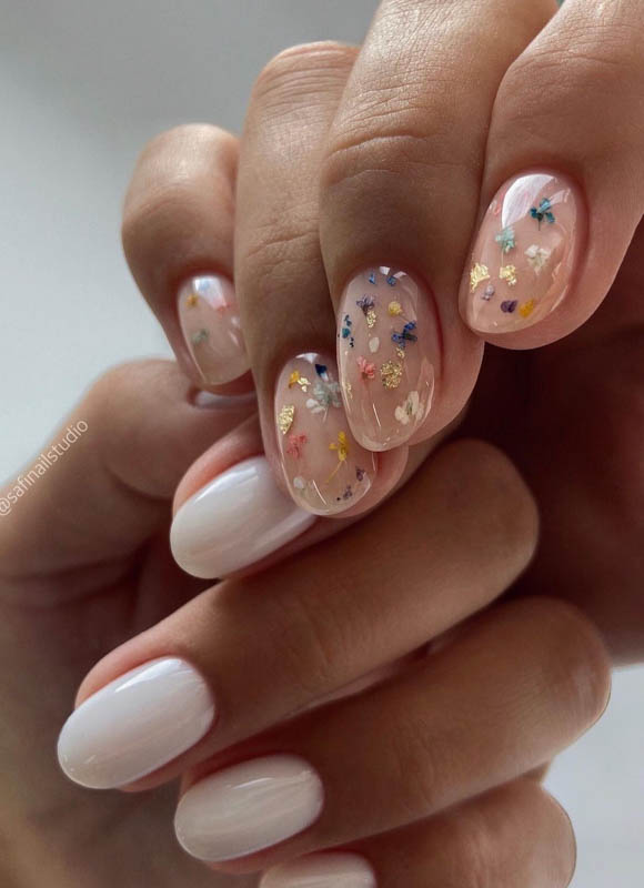 A close-up of a person's hands showcasing their elegant nail art, featuring clear polish adorned with delicate, colorful flowers and stars on some nails and a glossy white finish on the others.

