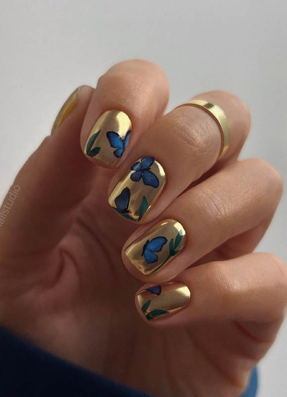 A hand with golden nails and blue floral and butterfly designs, wearing gold rings.

