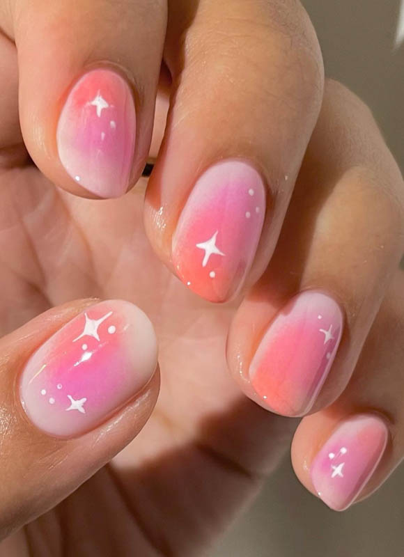 A hand with pink and white gradient nails and white star designs. Spring Nail Ideas For 2024.