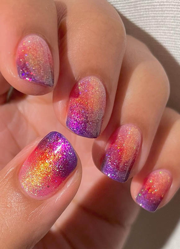 A hand with glitter gradient nails in purple, orange, yellow and red colors.
Spring Nail Ideas For 2024.