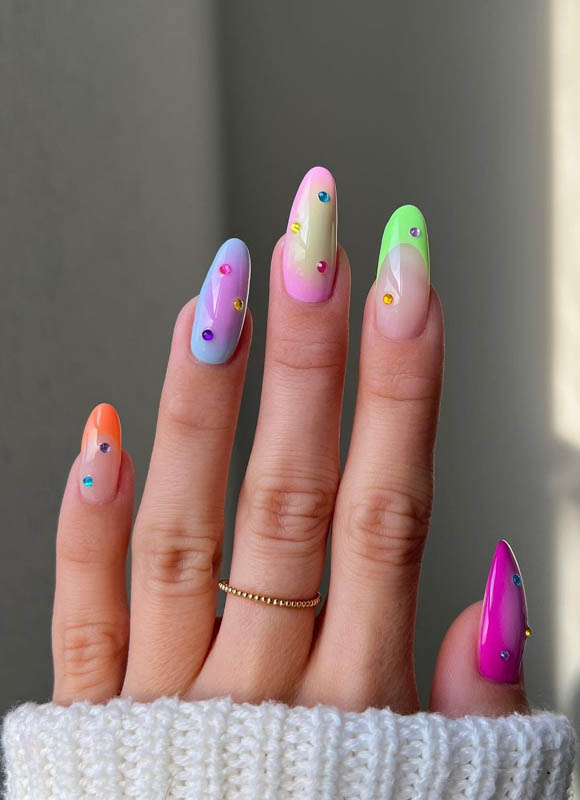 A hand with gradient colorful nails and jewels against a grey background

