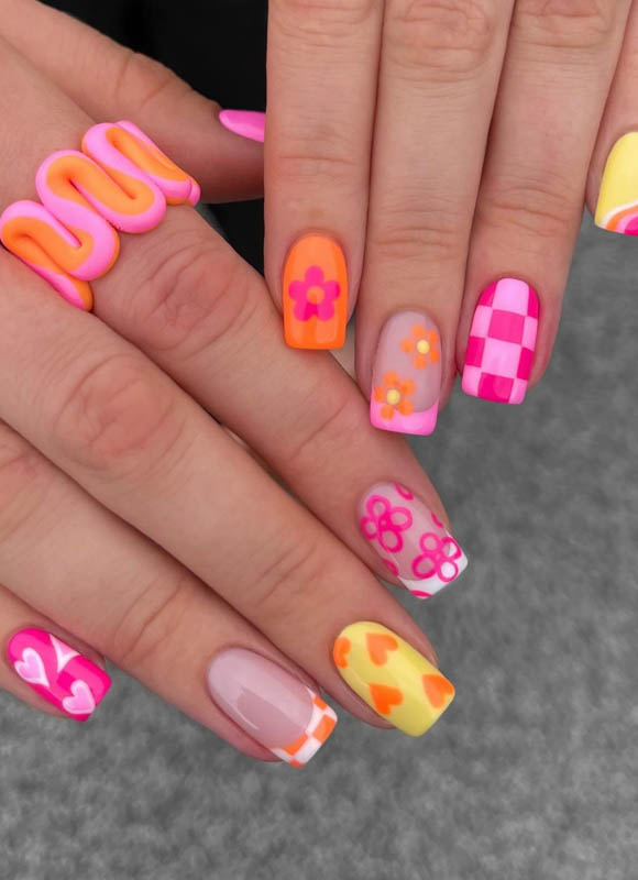 A close-up of hands showcasing a variety of brightly colored and creatively designed nail art, including patterns of hearts, flowers, and abstract designs in pink, orange and yeallow.

