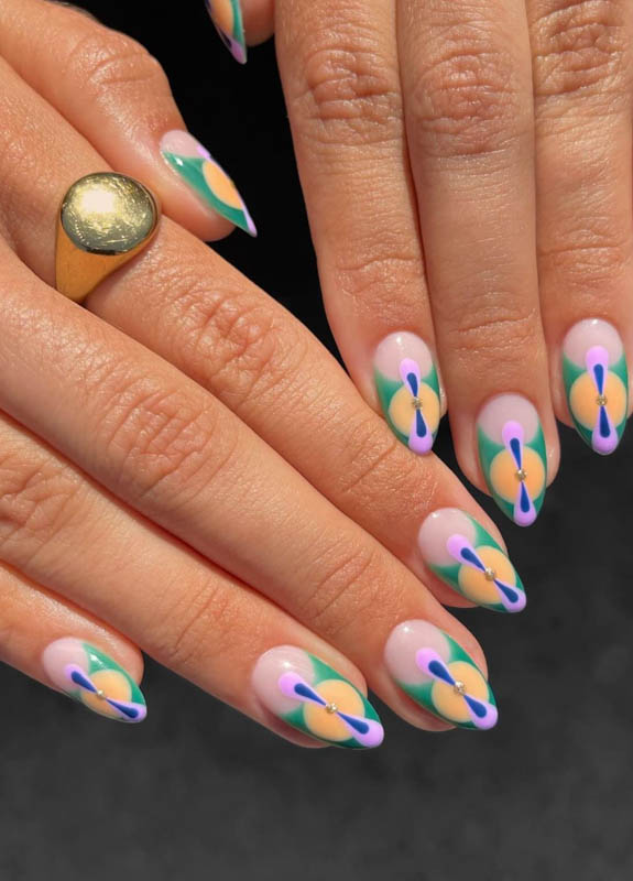 A close-up image of a hand adorned with a gold ring, showcasing long nails painted with colorful and artistic flower-like designs against a dark background.

